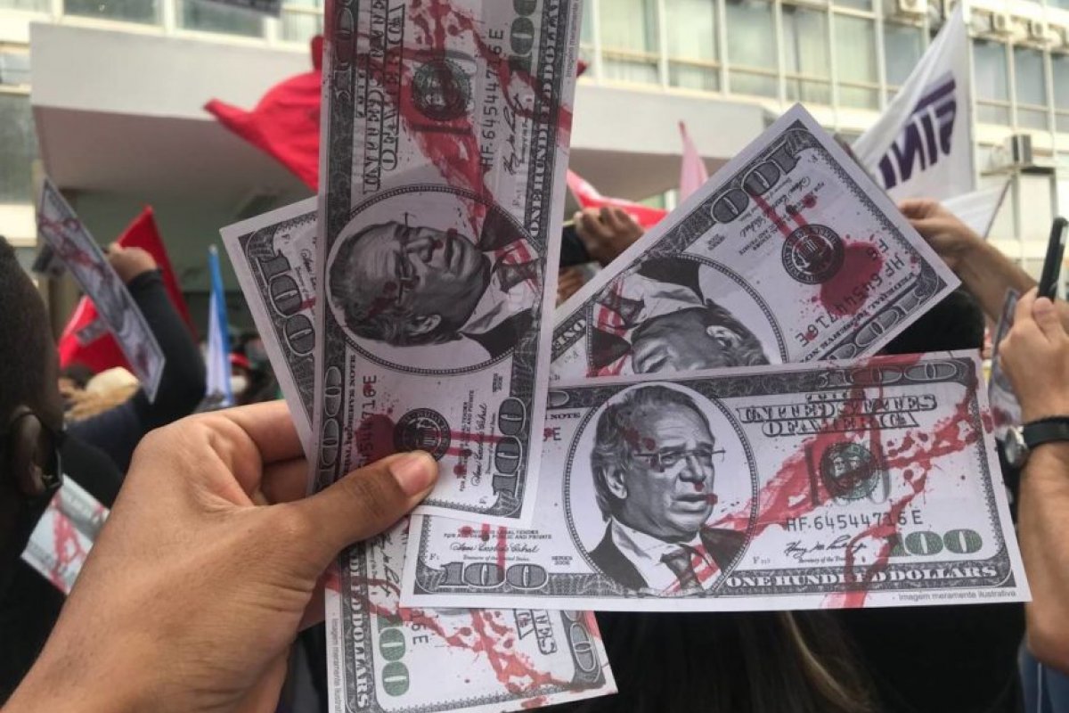 [Manifestantes jogam “dólares” com o rosto de Guedes em frente ao Ministério da Economia]